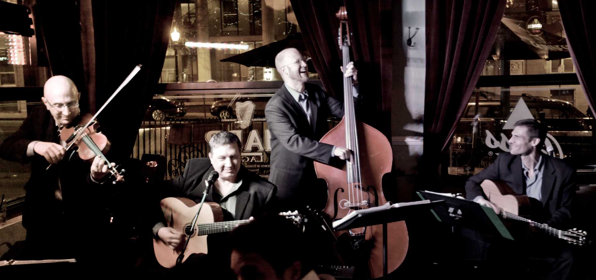 The band Gypsy Swing Revue performing live against a city backdrop, with violin, lead guitar, upright bass, and rhythm guitar.